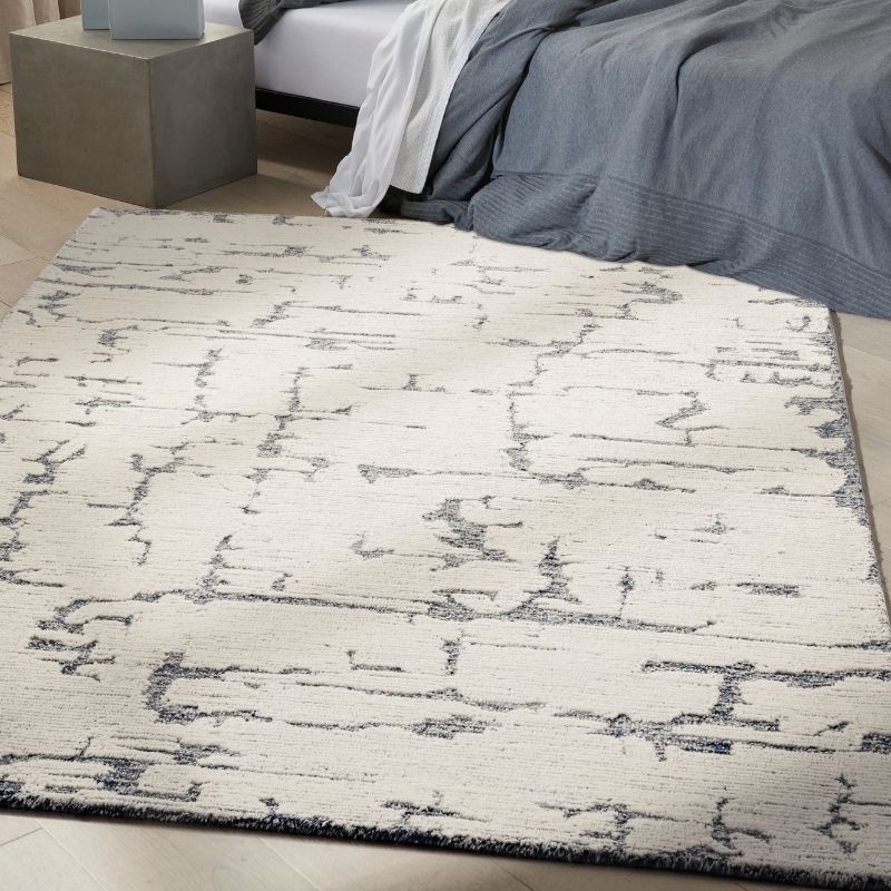 A rectangular area rug with an abstract distressed pattern in off-white and dark gray, styled in a modern bedroom setting next to a bed with gray bedding.