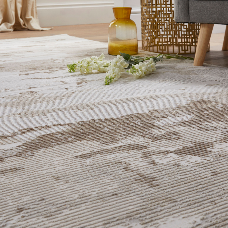 A close-up of a textured area rug with a distressed abstract pattern in beige and off-white, styled in a living room setting with a glass vase, white flowers, and a chair leg visible.