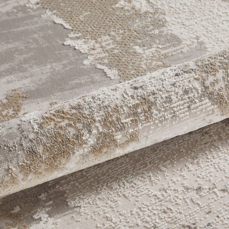 A close-up of a textured area rug with an abstract distressed pattern in beige, cream, and light gray tones, shown with a rolled section to highlight the pile and weave.
