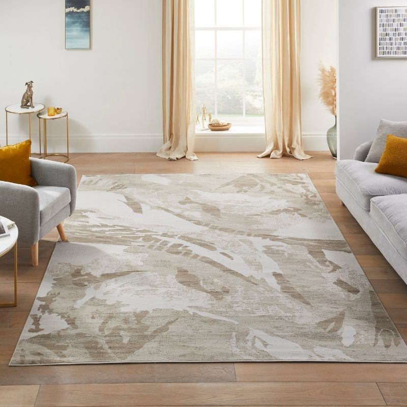 A large rectangular area rug with an abstract botanical pattern in beige, taupe, and off-white, styled in a bright modern living room with a gray sofa, armchair, and wood floors.