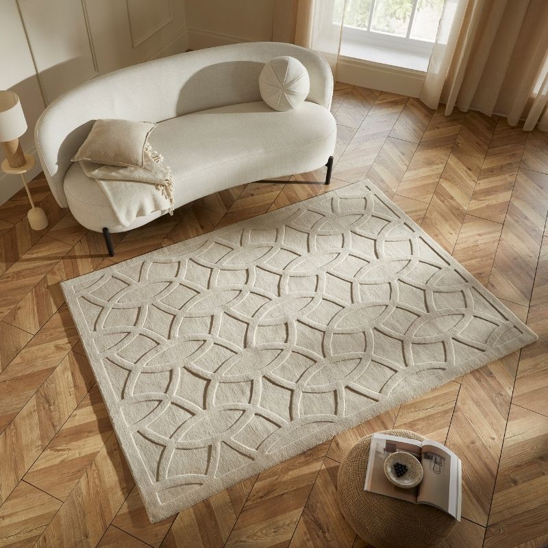 A large rectangular area rug with a raised geometric lattice pattern in cream and light beige, styled on a herringbone wood floor next to a curved white sofa.