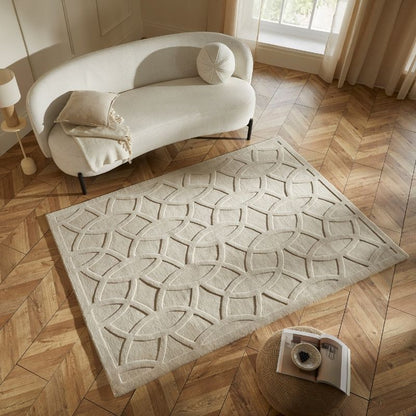 A large rectangular area rug with a raised geometric lattice pattern in cream and light beige, styled on a herringbone wood floor next to a curved white sofa.