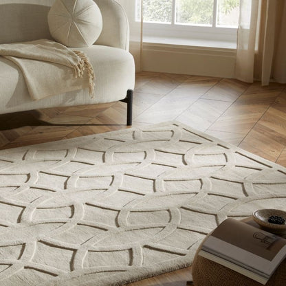 A cream-colored area rug with a raised geometric interlocking pattern, styled in a bright living room next to a white armchair and a wooden floor.