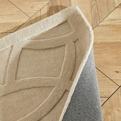 A close-up of a textured area rug with a raised geometric pattern in beige, showing its underside with a gray backing, placed on a light wood parquet floor.