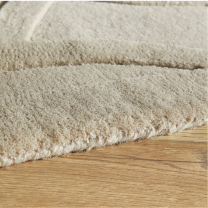 Close-up of a plush, textured area rug in a warm cream or off-white color, shown folded over to display its pile and edge, resting on a light brown wooden floor.