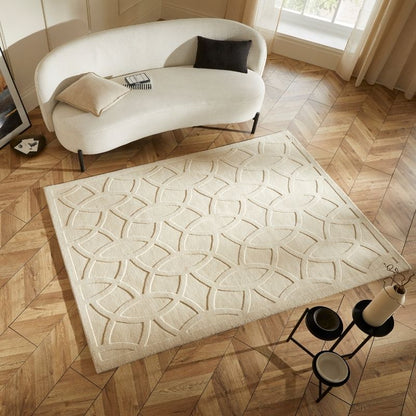 A cream-colored rectangular area rug with a raised geometric leaf pattern, styled on a light wood herringbone floor in a modern living room setting with a curved white sofa.