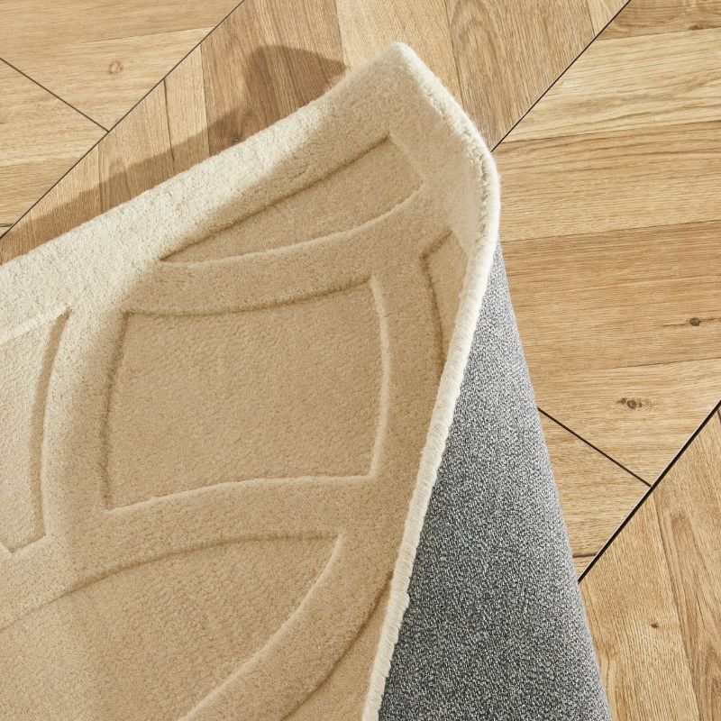A close-up of a folded area rug showing its beige patterned top and gray textured underside, placed on a light wood parquet floor.