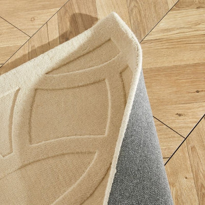 A close-up of a folded area rug showing its beige patterned top and gray textured underside, placed on a light wood parquet floor.