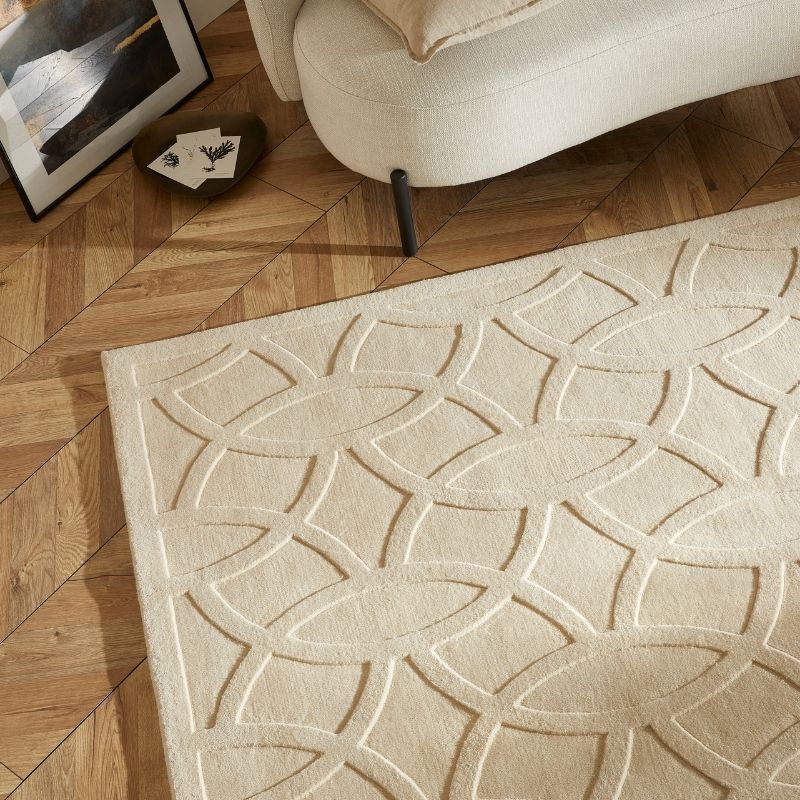 A cream-colored area rug with a raised geometric interlocking oval pattern, shown on a light brown wood parquet floor next to a modern cream sofa.