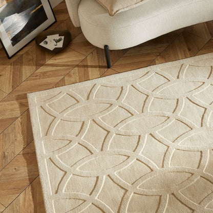 A cream-colored area rug with a raised geometric interlocking oval pattern, shown on a light brown wood parquet floor next to a modern cream sofa.