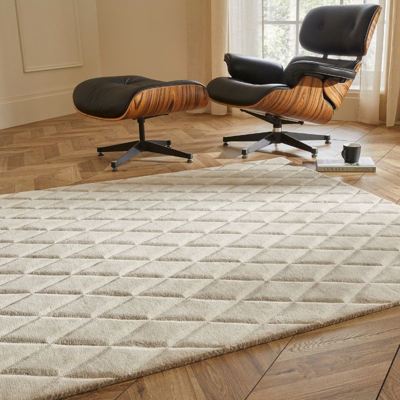 A large rectangular area rug with a raised diamond pattern in a light beige/cream color, shown in a styled living room setting with a wooden floor, an Eames-style lounge chair, and an ottoman.