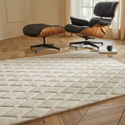 A large rectangular area rug with a raised diamond pattern in a light beige/cream color, shown in a styled living room setting with a wooden floor, an Eames-style lounge chair, and an ottoman.