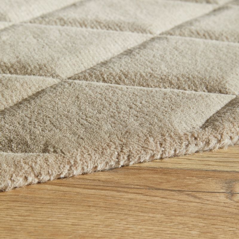 A close-up of a plush, textured area rug in a warm beige or cream color, featuring a raised geometric diamond pattern, shown partially folded over to reveal its soft pile and fringed edge on a light wood floor.