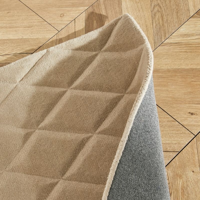 A close-up of a beige quilted rug with a gray underside and a cream stitched edge, shown partially folded over on a light wood parquet floor.