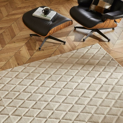 A large cream-colored area rug with a raised diamond quilted pattern, shown in a styled living room setting with two Eames-style lounge chairs and a wooden parquet floor.