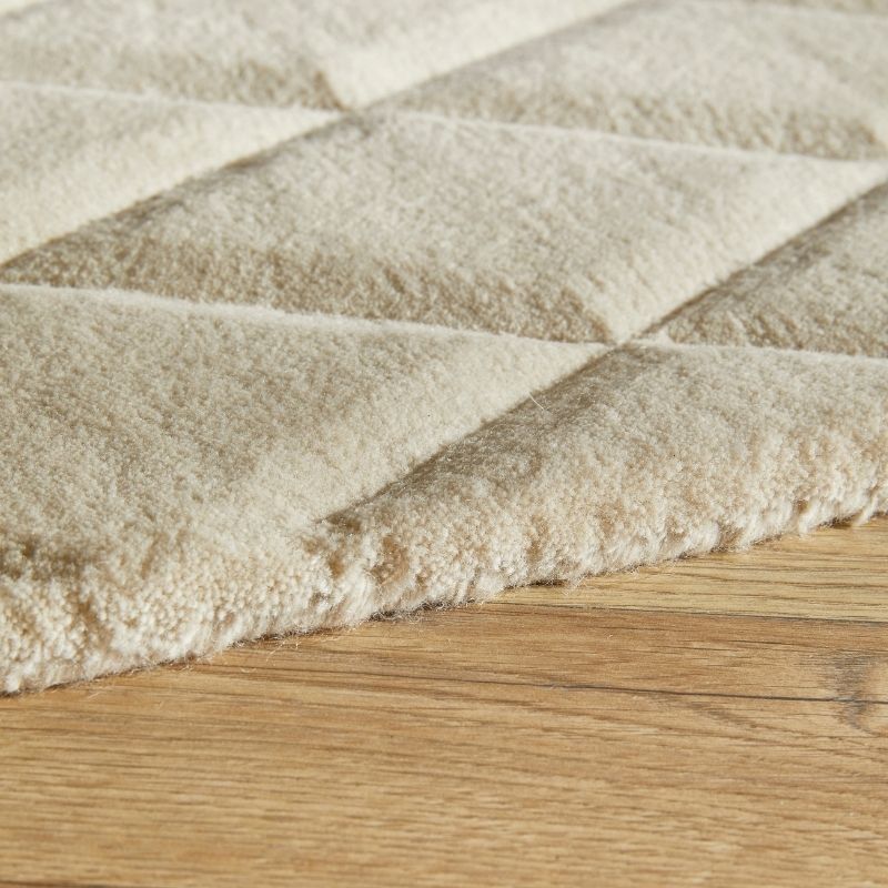 Close-up of a plush, diamond-quilted rug in a warm cream or beige color, shown resting on a light brown wooden floor.