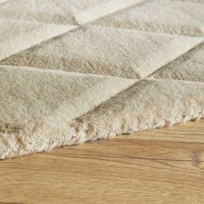 Close-up of a plush, diamond-quilted rug in a warm cream or beige color, shown resting on a light brown wooden floor.