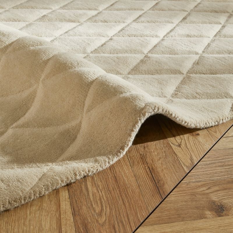 A close-up of a plush, quilted area rug in a warm cream or beige color, shown draped over a light brown wooden floor to display its soft texture and pile.