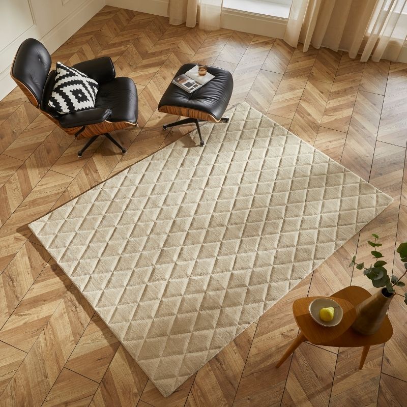 A large rectangular area rug with a diamond-quilted texture in a warm cream or light beige color, styled in a modern living room setting on a herringbone wood floor next to a black leather lounge chair and ottoman.