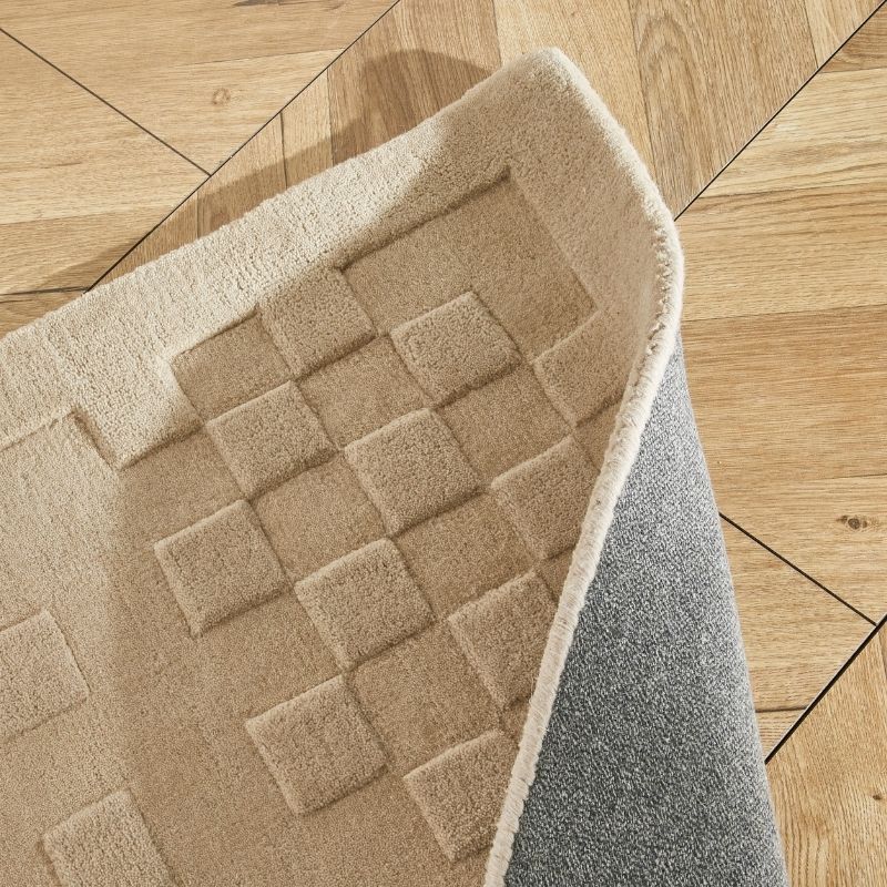 A close-up of a beige textured rug with a raised checkerboard pattern, shown partially folded over to reveal a gray backing, placed on a light wood floor.