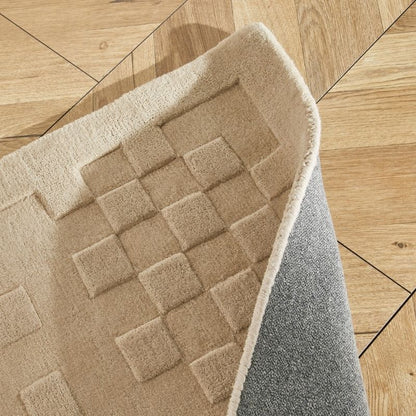 A close-up of a beige textured rug with a raised checkerboard pattern, shown partially folded over to reveal a gray backing, placed on a light wood floor.