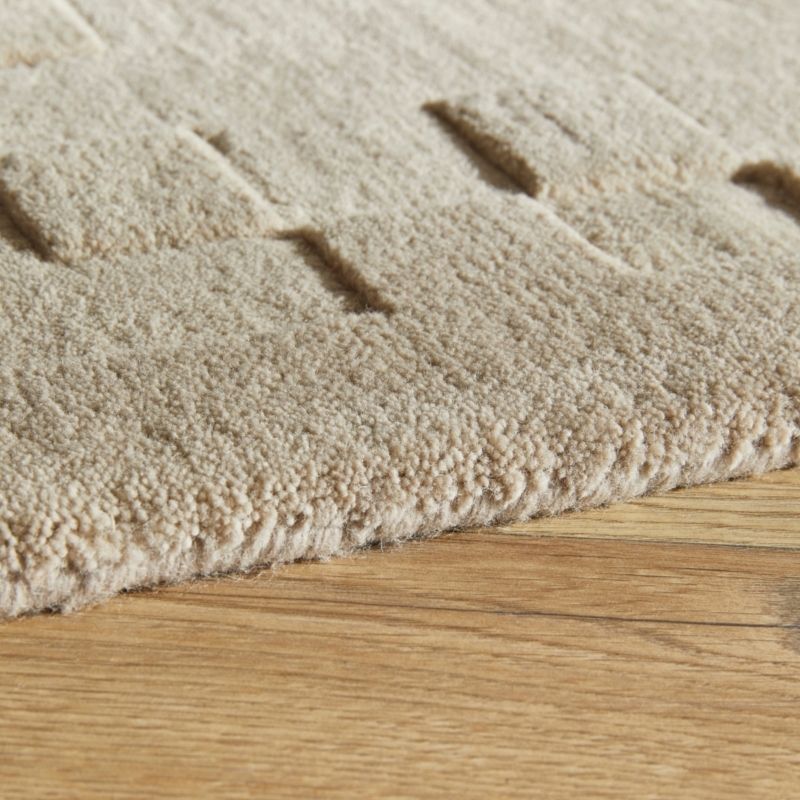 Close-up of a beige textured rug with a looped pile and subtle geometric channel tufting, shown resting on a light wood floor.
