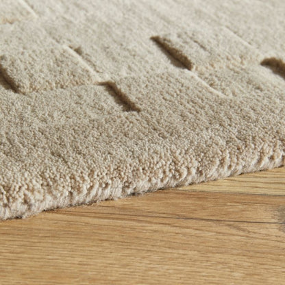 Close-up of a beige textured rug with a looped pile and subtle geometric channel tufting, shown resting on a light wood floor.