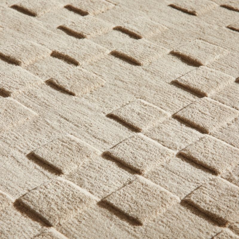 A close-up of a textured beige fabric or carpet with a repeating basketweave pattern in raised and recessed sections.