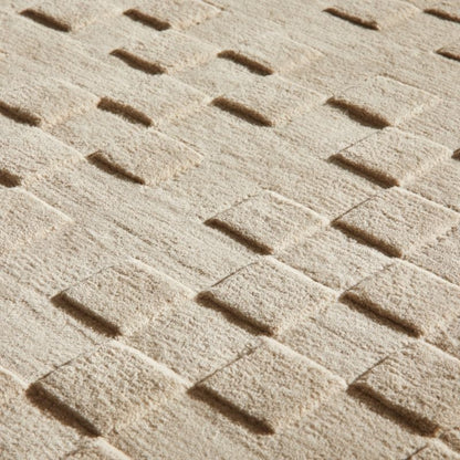 A close-up of a textured beige fabric or carpet with a repeating basketweave pattern in raised and recessed sections.