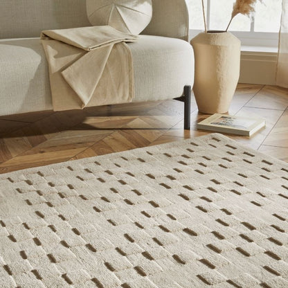 A cream-colored textured area rug with a geometric pattern of raised rectangular shapes, styled in a modern living room setting next to a beige sofa and a ceramic vase.