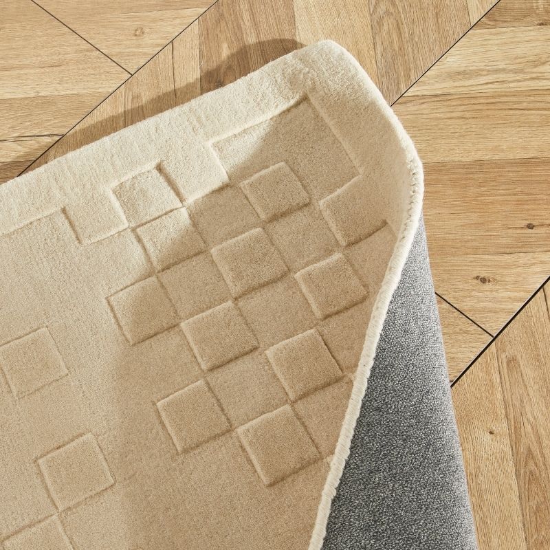 A close-up of a cream-colored area rug with a raised geometric grid pattern, shown partially curled over to reveal a gray backing, placed on a light wood parquet floor.