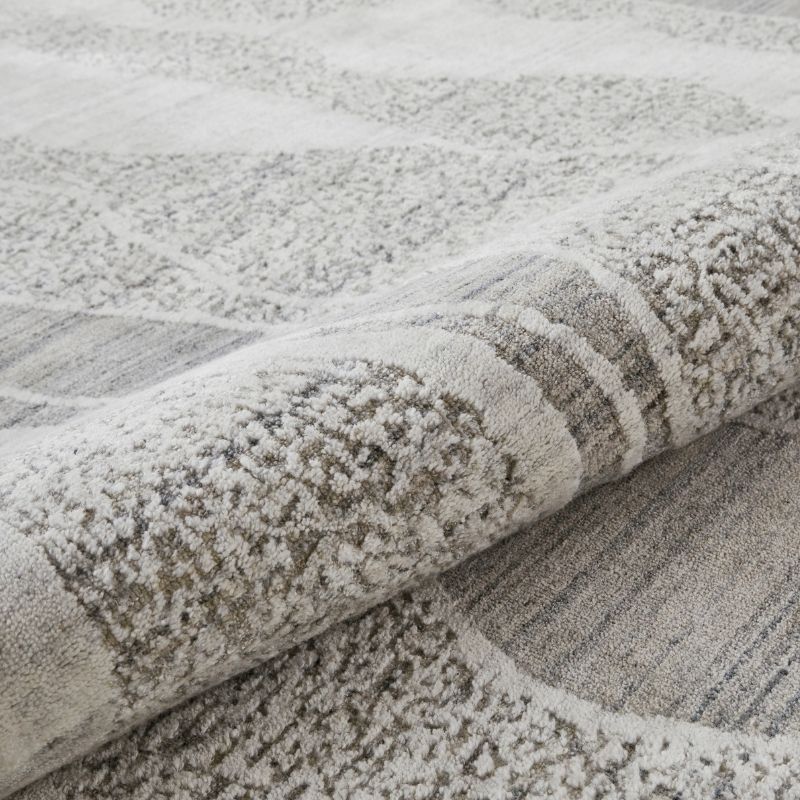 A close-up of a textured area rug with a modern abstract pattern in shades of gray, beige, and off-white, shown with one corner rolled up to reveal the pile.