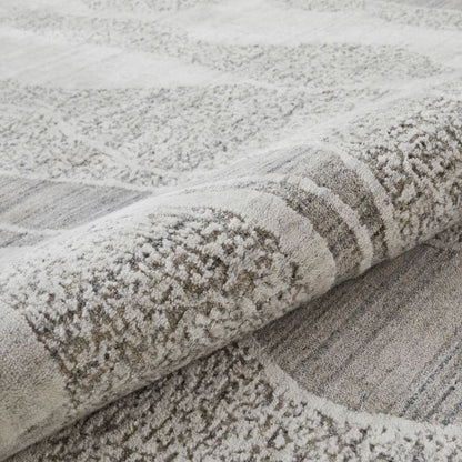 A close-up of a textured area rug with a modern abstract pattern in shades of gray, beige, and off-white, shown with one corner rolled up to reveal the pile.