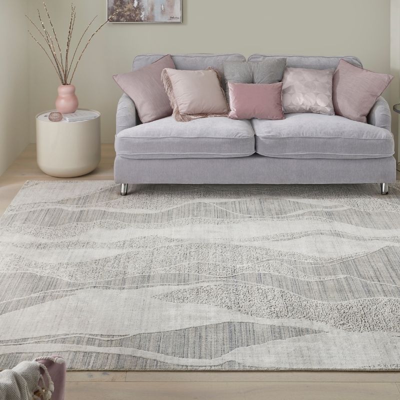 A light gray and white patterned area rug in a living room setting, styled under a light gray fabric sofa with pink and gray accent pillows, next to a round side table with a pink vase.