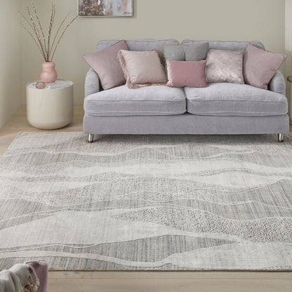 A light gray and white patterned area rug in a living room setting, styled under a light gray fabric sofa with pink and gray accent pillows, next to a round side table with a pink vase.
