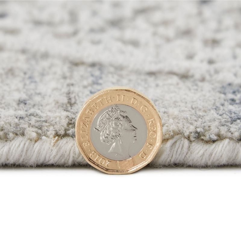 A close-up of a plush, textured area rug in a light gray and white pattern, with a British two-pound coin placed on top to show scale.