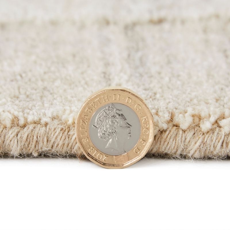 A close-up of a cream-colored textured rug with a fringed edge, shown with a British coin placed on top for scale against a white background.
