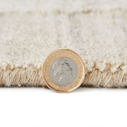 A close-up of a cream-colored textured rug with a fringed edge, shown with a British coin placed on top for scale against a white background.