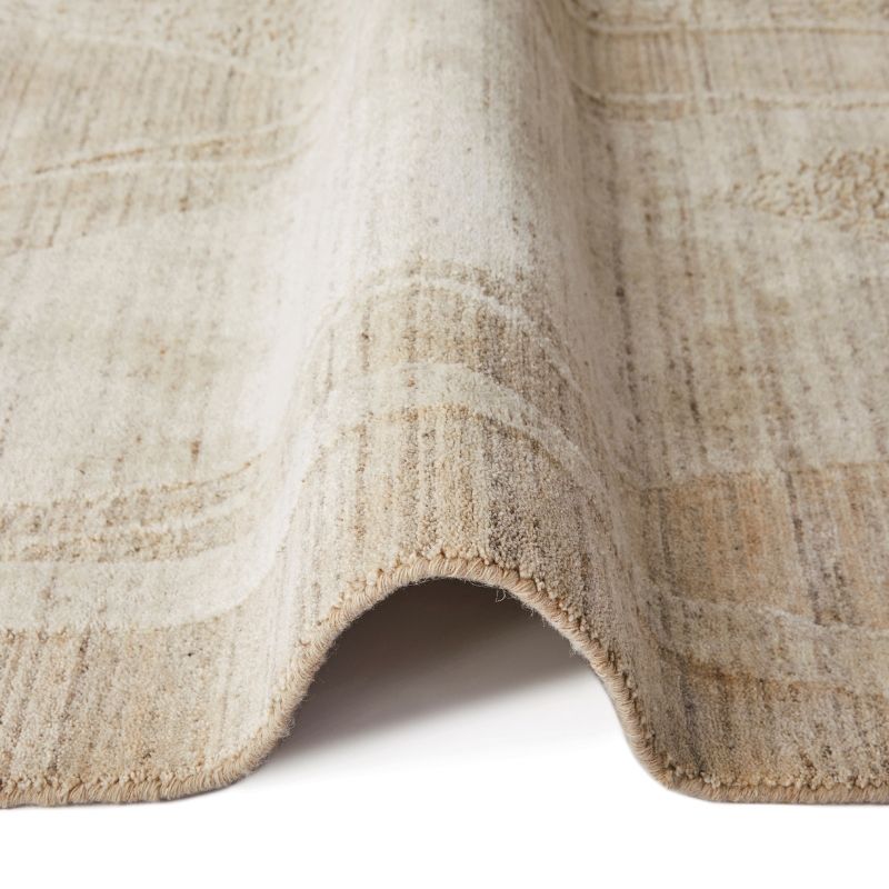 A close-up of a textured area rug with a distressed, abstract pattern in shades of beige, cream, and light brown, folded over to show its pile and edge.