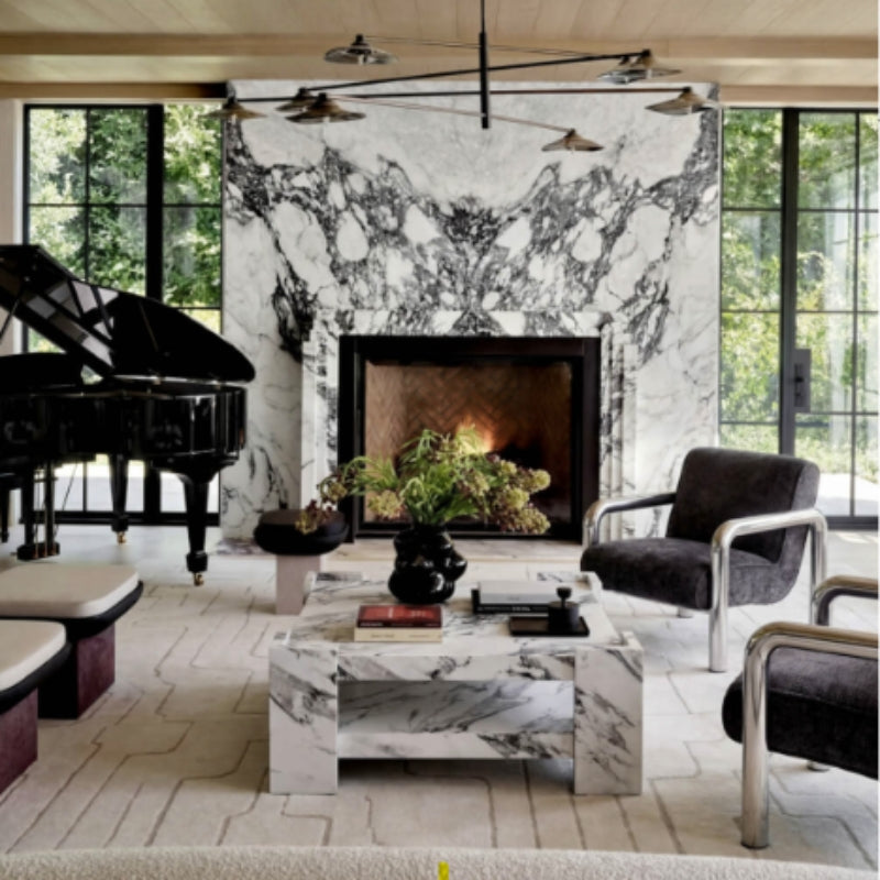 A luxury modern living room featuring a large black and white marble fireplace wall, a white marble coffee table, two dark gray armchairs with chrome frames, a black grand piano, and large black-framed windows.